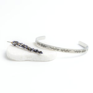 May include: A silver bangle bracelet with a delicate engraved design of leaves. The bracelet is displayed on a white stone with a dried lavender sprig.