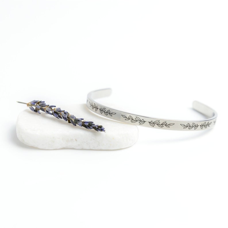 May include: A silver bangle bracelet with a delicate engraved design of leaves. The bracelet is displayed on a white stone with a dried lavender sprig.