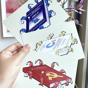 May include: Three art prints showing cartoon creatures around books. The books are titled "DSM-5", "The C Programming Language", and "The Communist Manifesto". The creatures are blue, yellow, and red, with white flower accents.