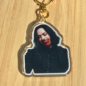 May include: A clear acrylic keychain featuring a smiling person with long dark hair and blood around their mouth. The keychain has a gold-colored heart-shaped clasp.