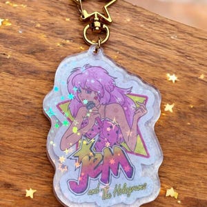May include: A clear acrylic keychain featuring a cartoon illustration of a woman with pink hair holding a microphone. The keychain has a gold star-shaped clasp and the text "JEM and The Holograms". The design includes holographic accents.