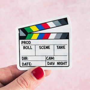 May include: A white film clapperboard with black, gray, yellow, red, and blue accents. The clapperboard has the words "PROD," "ROLL," "SCENE," "TAKE," "DIR," "CAM," "DATE," and "DAY NIGHT" printed on it. The clapperboard is held in a hand with red nail polish.
