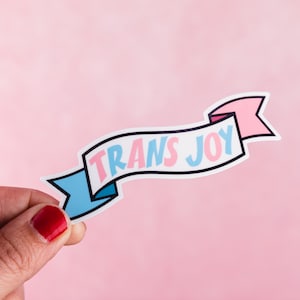 May include: A pink and blue banner with the text "TRANS JOY" in white letters. The banner is held by a hand with a red nail polish.