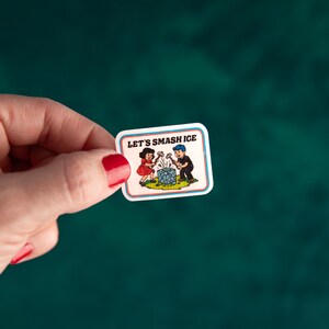May include: A rectangular sticker with a white border and the text "LET'S SMASH ICE." The sticker features a cartoon illustration of a girl and a boy smashing a blue ice cube with hammers. The background is a teal green.
