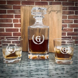 May include: A personalized whiskey decanter set with two glasses. The decanter and glasses are etched with the letter "G" and a laurel wreath. The set is presented in a wooden box with the name "George" inscribed on the front.