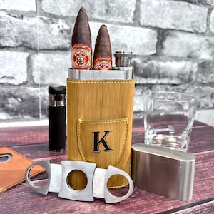 May include: A cigar set featuring two cigars, a lighter, a cigar cutter, and a flask in a tan leather case with the letter "K". A small glass and a metal container are also present. The cigars have red and gold labels.