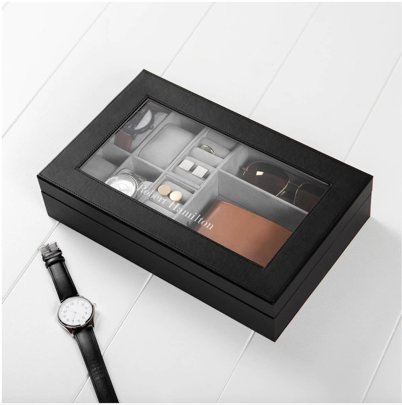 May include: Black leather watch box with a clear lid. The box has compartments for watches, cufflinks, and sunglasses. The box is personalized with the name "Robert Hamilton".