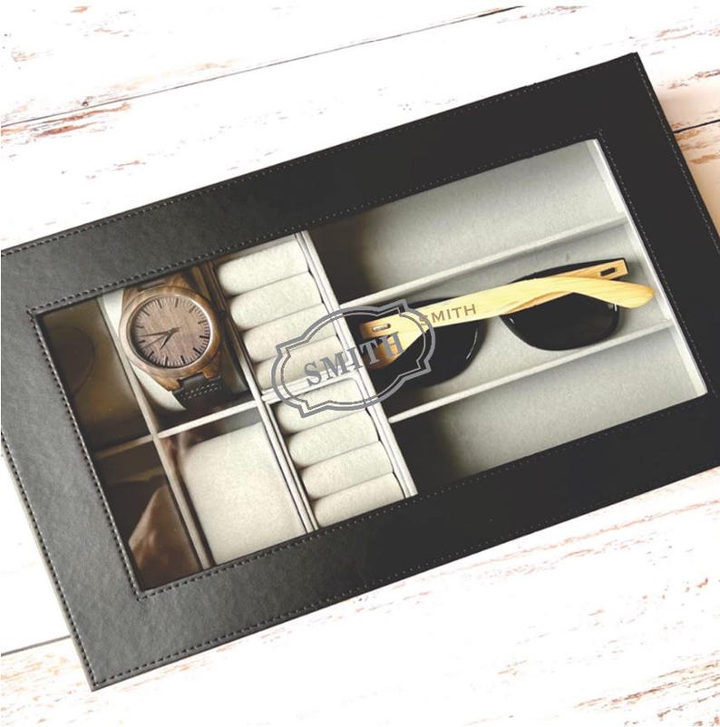 May include: Black leather watch and sunglasses box with a clear lid. The box has compartments for watches, rings, and sunglasses. The box is personalized with the name "SMITH".