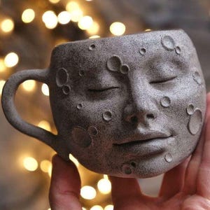May include: A ceramic mug shaped like a face with closed eyes and a subtle smile. The mug is decorated with craters, resembling the surface of the moon.