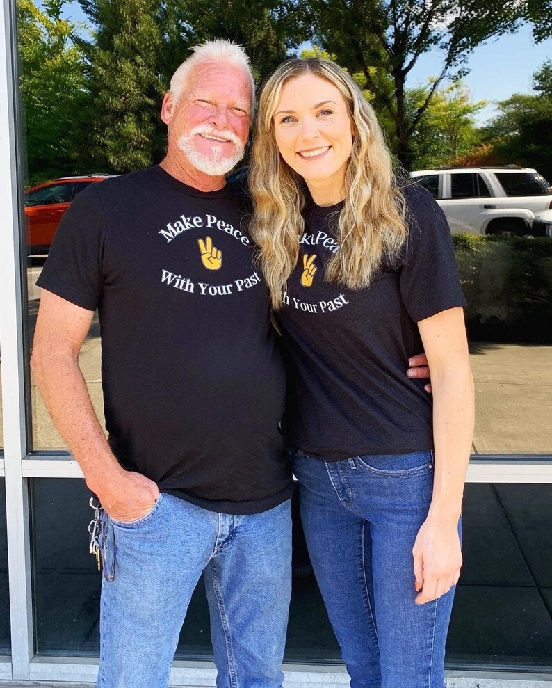 May include: Two people wearing black t-shirts with the text "Make Peace With Your Past" and a peace sign graphic. The person on the left is wearing blue jeans and the person on the right is wearing blue jeans.