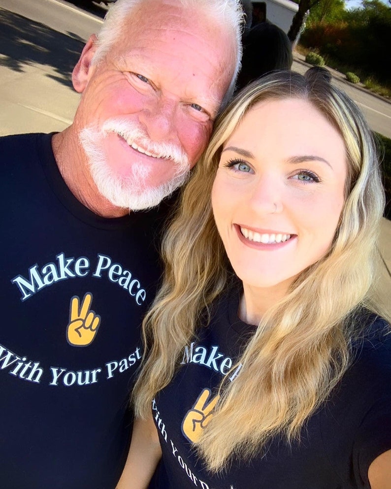 May include: A father and daughter wearing black t-shirts with the text "Make Peace With Your Past" and a peace sign graphic. The shirts are black with white and yellow text and graphics.