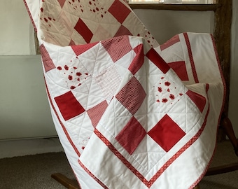 May include: A white and red patchwork quilt with a red and white gingham pattern. The quilt is folded and draped over a rocking chair.