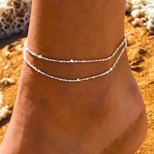 May include: A double-strand silver anklet with fine chain links. The anklet is worn on a foot on a sandy beach, with the sea in the background. A simple, elegant piece of jewellery.