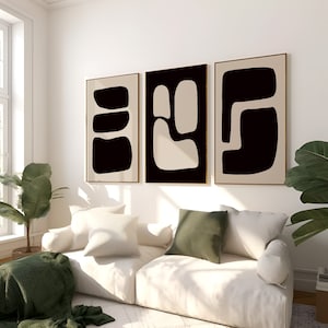 May include: Three framed abstract art prints. Each print features a unique design with black and beige organic shapes against a neutral background. The frames have a gold finish. The artwork hangs above a white sofa with pillows and a green blanket.
