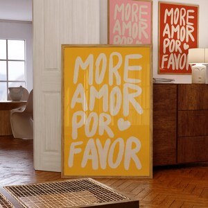 以下が含まれることがあります： 「MORE AMOR POR FAVOR」の白い文字とハートマークが入った、額縁入りの黄色のポスター。同様のテキストが書かれたピンクと赤のポスターも見える。ポスターは木製の床と家具のある部屋に飾られている。
