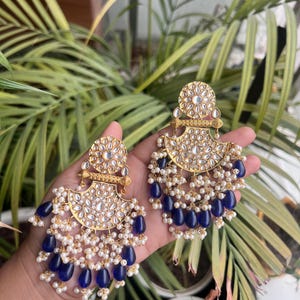 以下が含まれることがあります： 精巧なディテールが施された、ゴールドトーンのイヤリングのペア。イヤリングは、花の模様の円形トップ、透明な石の中央部分、青い涙型のビーズと小さな白い真珠で飾られた扇形のボトムが特徴です。