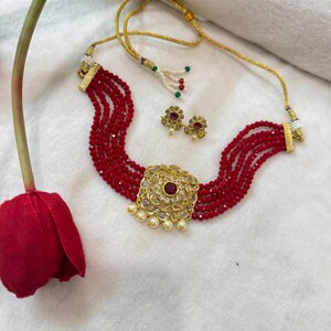 May include: A red beaded choker necklace with a gold pendant featuring a large red stone surrounded by smaller clear stones. The necklace is paired with matching red stone earrings.