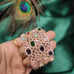 Pode incluir: Um conjunto de cinco pulseiras de ouro com pedras transparentes e coloridas. As pulseiras são decoradas com desenhos florais e apresentam pedras de forma retangular em tons de rosa, verde, azul e vermelho.