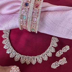 May include: A set of rose gold jewelry featuring a necklace, earrings, a ring, and three bangles. The jewelry is adorned with clear cubic zirconia stones and colorful gemstones.
