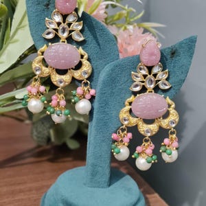 Peut inclure: Une paire de boucles d'oreilles dorées ornées de pierres roses. Les boucles d'oreilles présentent une pierre en forme de goutte d'eau en haut, une pierre ovale au centre et des grappes pendantes de perles roses, vertes et blanches. Elles sont exposées sur un présentoir en velours bleu sarcelle.