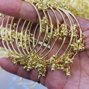 May include: A set of gold-coloured bangles. Each bangle features small, spherical decorations. The bangles are held in a hand, highlighting their detailed design and reflective surface. The jewellery appears to be made of a shiny, metallic material.