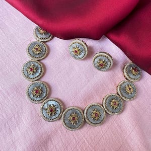 Puede incluir: Un collar de plata y oro con una serie de colgantes redondos que presentan un diseño blanco y gris con una piedra preciosa roja en el centro. El collar se combina con pendientes a juego.