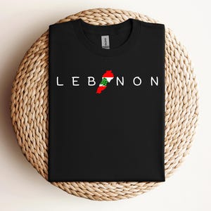 May include: Black t-shirt with the word "LEBANON" printed in white letters. The outline of Lebanon is in the center of the word, filled with the Lebanese flag colors of red, white, and green.