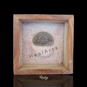 以下が含まれることがあります： ガラスの蓋が付いた木製の箱の中に、ピンクの砂とアメリカの25セント硬貨が少量入っています。砂には「HealArts」という文字が印刷されています。箱は黒い表面の上に置かれており、箱の下には「Ruby」という文字が印刷されています。