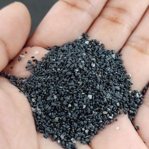 May include: A handful of dark gray, small, granular material, possibly sand or gravel, is held in an open palm. The material has a rough texture with small, light-colored particles mixed in. The image is a close-up, highlighting the texture and color of the material.