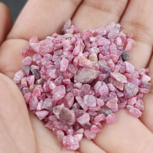 Könnte beinhalten: Eine Handvoll kleiner, grob geschnittener rosa Edelsteine. Die Steine variieren in der Farbe von hell- bis tiefrosa, wobei einige Anklänge von Grau und Braun aufweisen. Die Edelsteine sind unregelmäßig geformt und scheinen rohe, natürliche Kristalle zu sein.