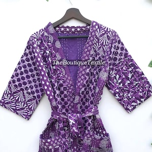 May include: Purple cotton robe with a white floral and polka dot pattern. The robe has a tie belt and two pockets. The text "TheBoutiqueTextile" is visible on the robe.