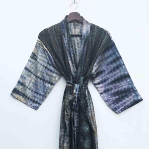 May include: A long, tie-dye robe in shades of black, gray, and blue hangs against a white background. The robe has wide sleeves and a matching belt. The fabric appears soft and flowing, with a subtle sheen.