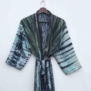 May include: A tie-dye patterned robe in shades of blue, green, and black hangs on a wooden hanger. The robe features a belt and a flowing design, with a mix of striped and patterned sections.