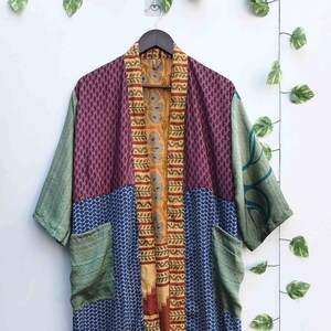 May include: A patchwork kimono robe with a variety of patterns and colors. The robe features a mix of purple, blue, green, and gold fabrics. It has short sleeves and two pockets. The robe is hanging on a wooden hanger against a white wall.