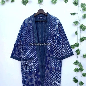 May include: A blue and white kantha quilted cotton robe with a patchwork design. The robe has a long, open front and three-quarter sleeves. The robe is hanging on a wooden hanger against a white background.