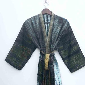 May include: A tie-dye patterned robe in shades of black, green, and blue hangs on a wooden hanger. The robe features a matching belt and a textured design. The garment is displayed against a plain white background.