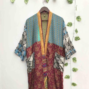 Może przedstawiać: Szlafrok typu kimono z długim rękawem o patchworkowym wzorze z różnymi motywami. Odzież zawiera ciemnoczerwoną sekcję z wzorem paisley, niebieską sekcję geometryczną oraz czarno-białe motywy wachlarzy na rękawach i kieszeniach, ze złotym wykończeniem.