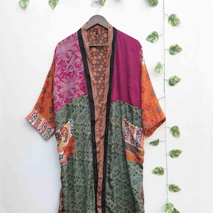 Puede incluir: Una bata estilo kimono de manga larga con un diseño de patchwork de varios patrones florales y de cachemira en tonos magenta, naranja y verde oliva. La prenda tiene un borde negro a lo largo de la abertura frontal y dos bolsillos.