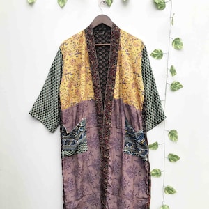 May include: A long-sleeved kimono-style robe featuring a patchwork design with yellow, purple, and dark green fabric panels. The garment includes two front pockets, a dark patterned trim along the collar, and various geometric and floral prints.
