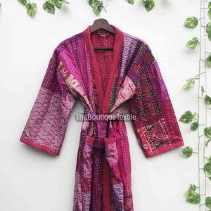 May include: A patchwork robe in shades of pink, purple, and red, with a matching belt. The robe features a quilted design and is hanging on a wooden hanger. The text "TheBoutiqueTextile" is visible on the robe.