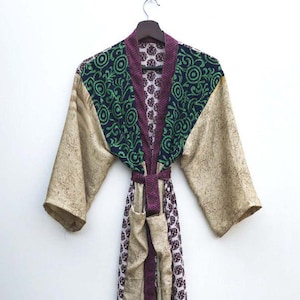 May include: A long, patterned robe with a belt, hanging against a white background. The robe features a mix of colors and patterns, including beige, dark green, and purple. The sleeves and body have different designs, and the robe is made of a soft, flowing fabric.