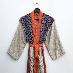 May include: A colorful, patterned robe with a paisley design on the sleeves and a blue and gold floral pattern on the collar. The robe has an orange trim and a matching belt. The robe is hanging on a wooden hanger.
