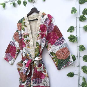 May include: A white cotton robe featuring a vibrant floral pattern in shades of pink, orange, and green. The garment includes long sleeves, a matching tie belt, two front pockets, and visible kantha stitching throughout. The text boutique textile is visible.