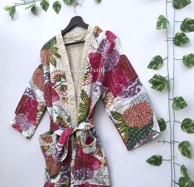 May include: A white cotton robe featuring a vibrant floral pattern in shades of pink, orange, and green. The garment includes long sleeves, a matching tie belt, two front pockets, and visible kantha stitching throughout. The text boutique textile is visible.