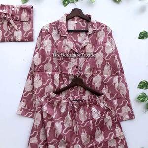 May include: A pink pajama set with a white cat print. The top has long sleeves and a button-up front. The pants have an elastic waistband.