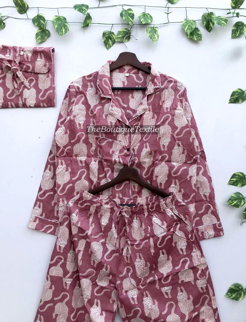 May include: A pink pajama set with a white cat print. The top has long sleeves and a button-up front. The pants have an elastic waistband.