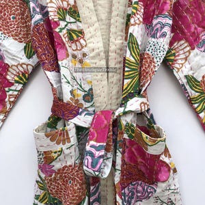 May include: A quilted robe featuring a white base with a vibrant floral pattern in shades of pink, orange, green, and yellow. The garment includes a beige collar, a matching tie belt, and two front pockets. The text TheBoutiqueTextile is visible.
