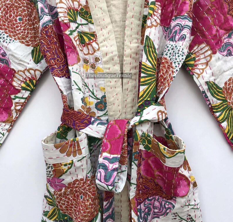 May include: A quilted robe featuring a white base with a vibrant floral pattern in shades of pink, orange, green, and yellow. The garment includes a beige collar, a matching tie belt, and two front pockets. The text TheBoutiqueTextile is visible.