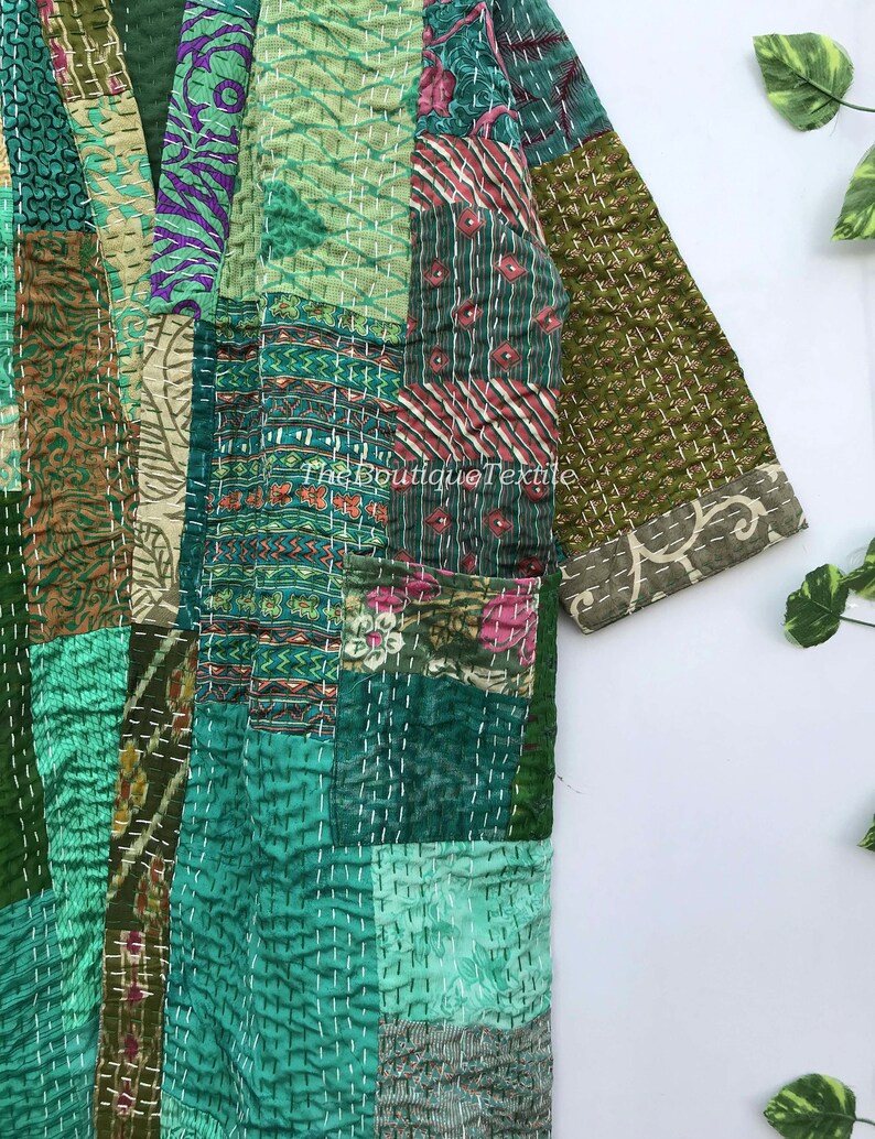 May include: A green patchwork kantha quilt jacket with a floral print. The jacket has a long sleeve and a button closure. The fabric is quilted with white thread.