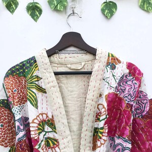May include: A quilted jacket featuring a white base with vibrant floral and abstract patterns in pink, orange, green, and yellow. The garment has a beige collar and front trim with visible red running stitch details and the text TheBoutiqueTextile.